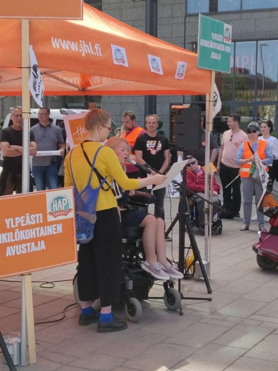 Kuvassa Hetan varapuheenjohtaja Elina Nieminen pitää puhetta Henkilökohtaisen avun paraatin alkutilaisuudessa. Hän on oranssin teltan edustalla, telttaa ympäröivät paraatin logot. Taustalla näkyy yleisöä.