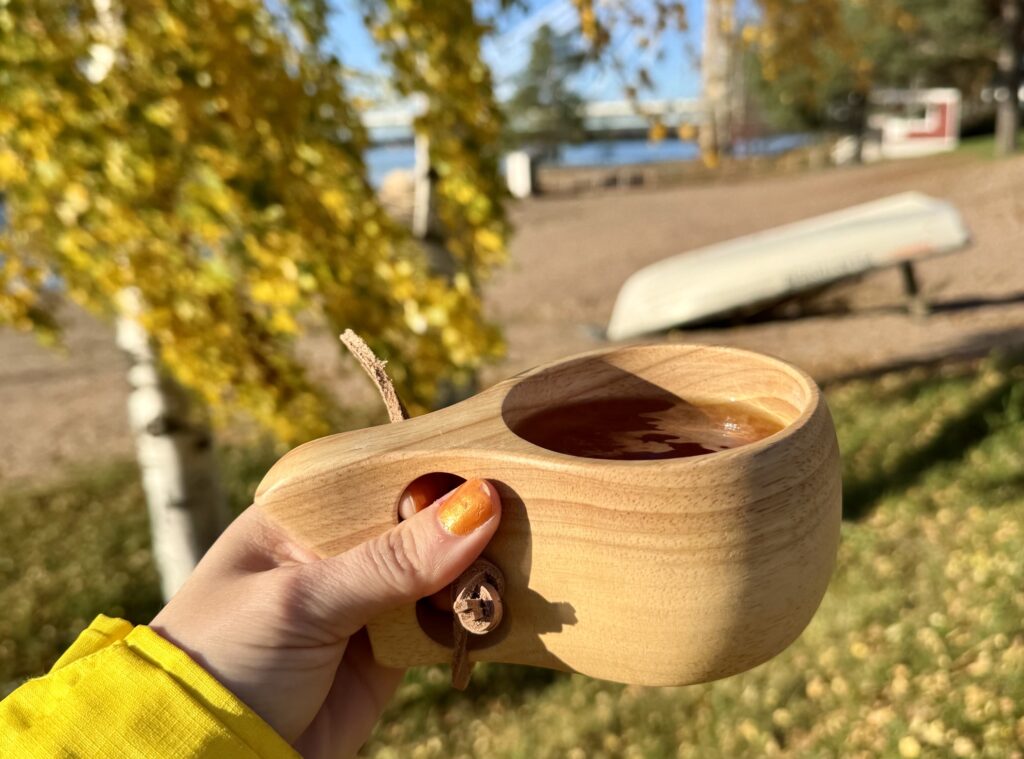 henkilön kädessä kuksa, taustalla syksyistä joenrantamaisemaa.