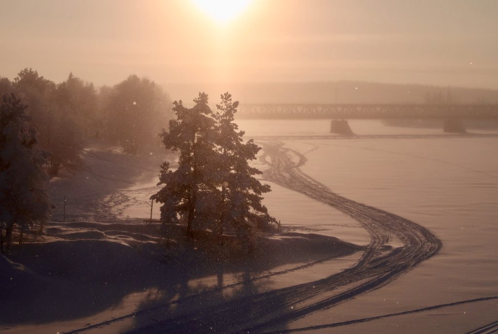 Talvinen, luminen maisema auringonlaskussa. Järven jäätä, puita ja silta. 