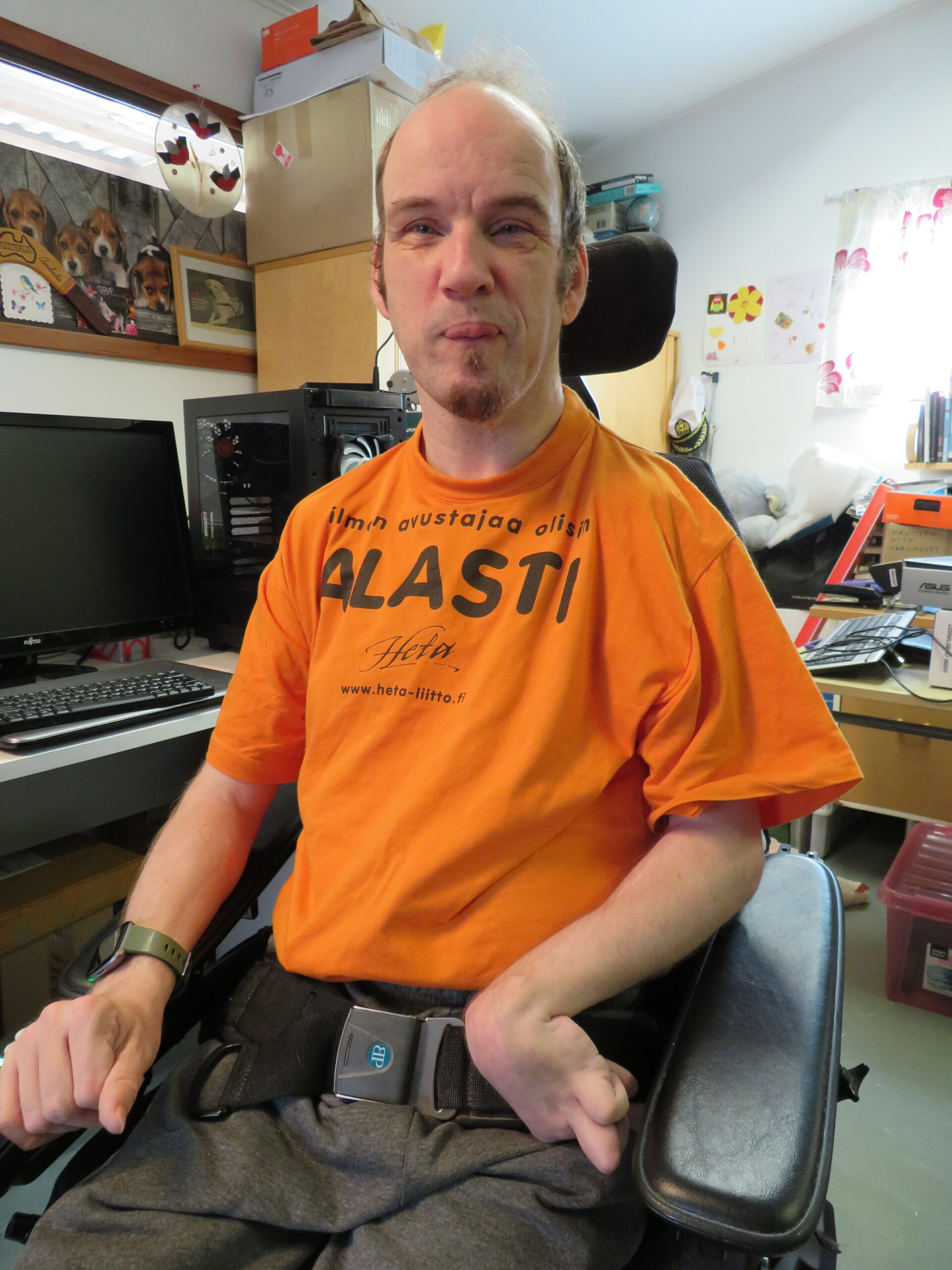 Jan Hänninen istuu pyörätuolissaan oranssissa T-paidassa, jossa lukee: Ilman avustajaa olisin alasti.