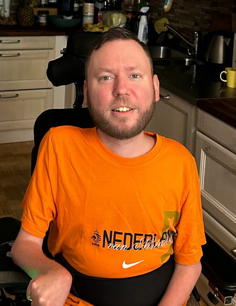 Lassi Murto istuu pyörätuolissa keittiössä. Hänellä on yllään oranssi T-paita. 