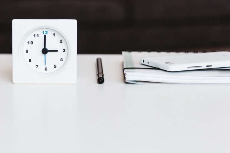 Kuvituskuva, jossa on valkoinen neliönmuotoinen kello, kynä ja kalenteri valkoisella pöydällä.