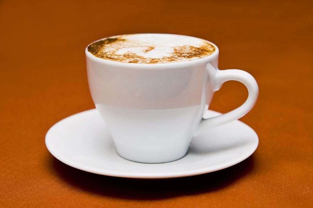 VertaisKahvit työnantajille -sivun kuva. Valkoinen kahvikuppi, jossa on maitokahvia. Kuvan tausta on syvän oranssi.