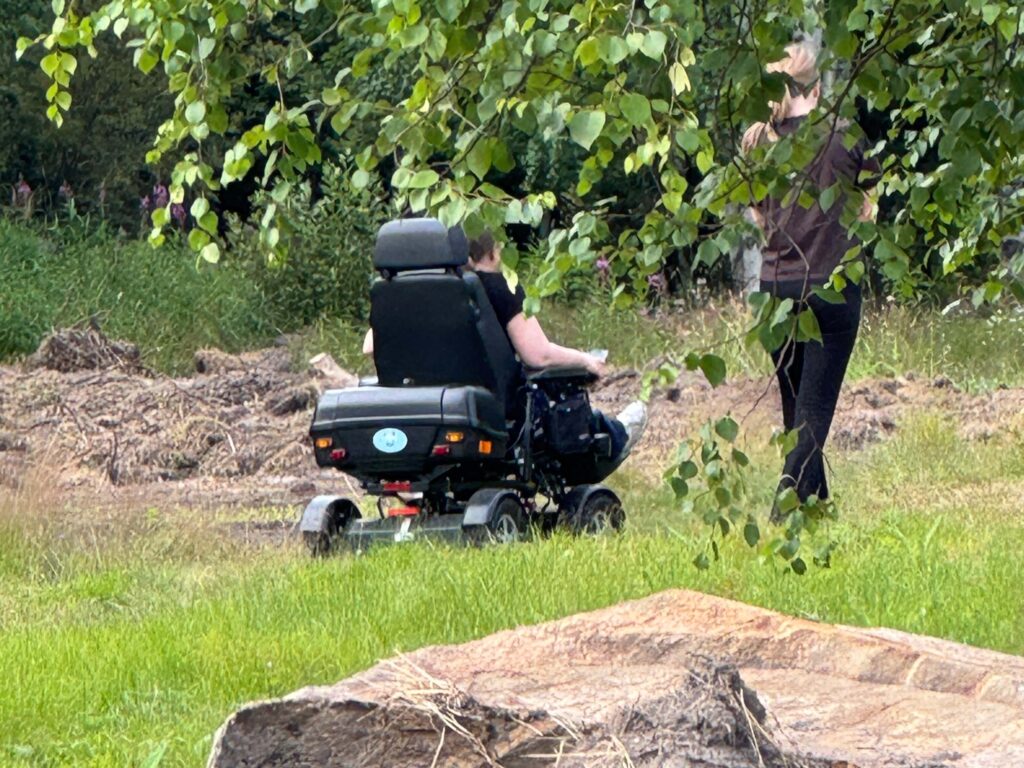 Nainen ajaa sähköpyörätuolilla luonnossa. Vieressä kävelee avustaja. Etu-alalla iso kivi ja koivun oksia.
