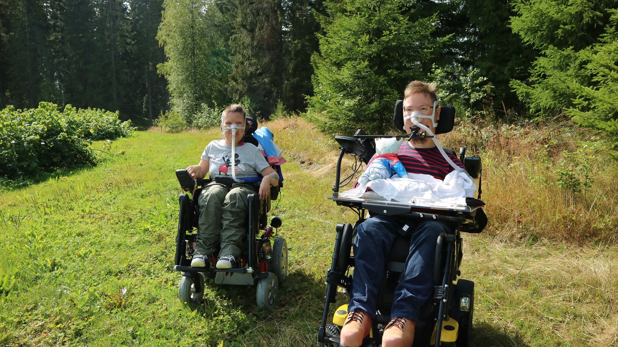 Pietari ja Maiju, jotka istuvat pyörätuoleissaan. Taustalla näkyy vihreä, kesäinen maisema.